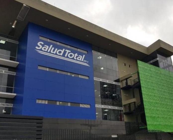 Fachada institucional de una sede de Salud Total EPS en Colombia, con pacientes siendo atendidos en la recepción por personal de salud uniformado