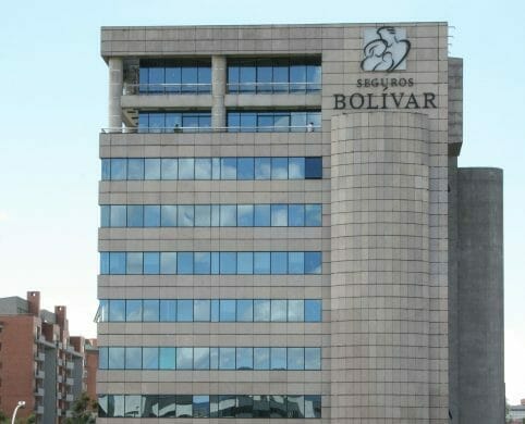 Asesor de Seguros Bolívar atendiendo a un cliente colombiano en una oficina comercial con el logo de la aseguradora visible al fondo